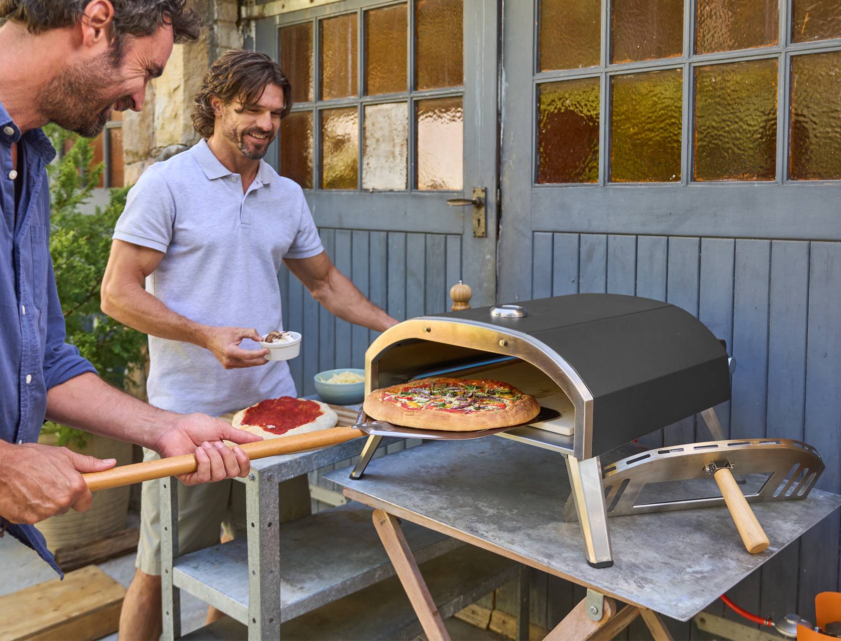 Két férfi pizzát süt egy kerti pizzasütőben, az egyik éppen beteszi a pizzát a sütőbe.