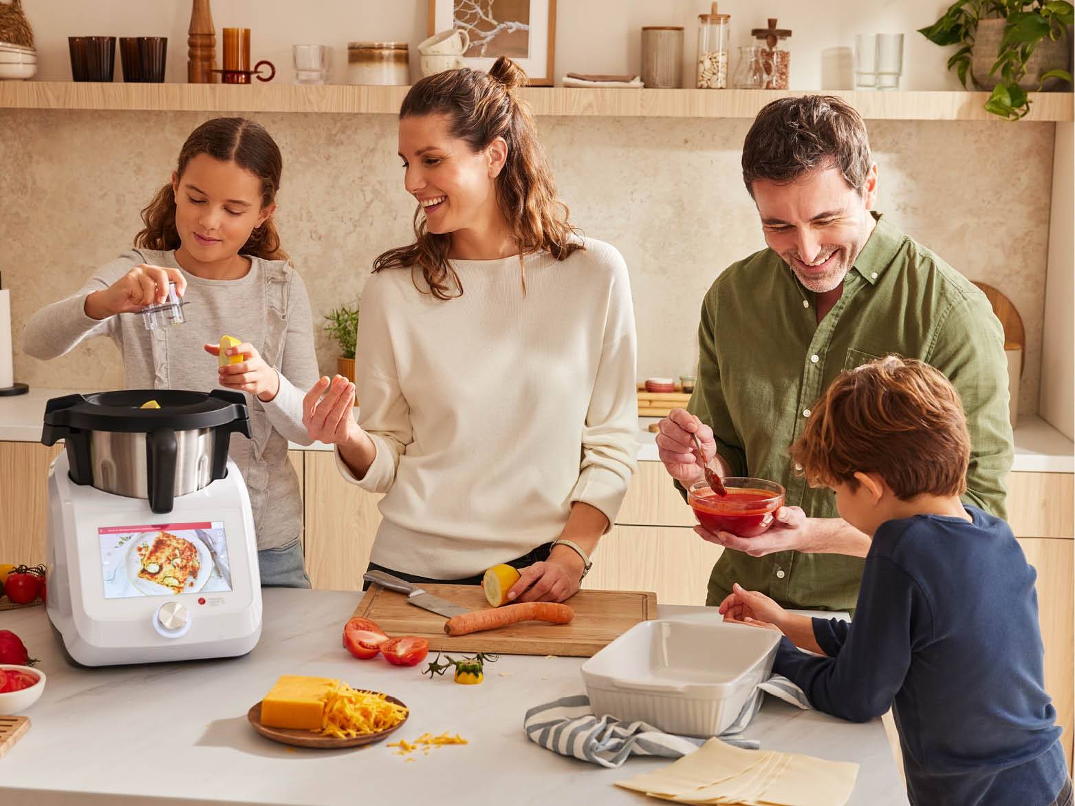 Család főz a konyhában, robotgéppel, zöldségekkel és sajttal.