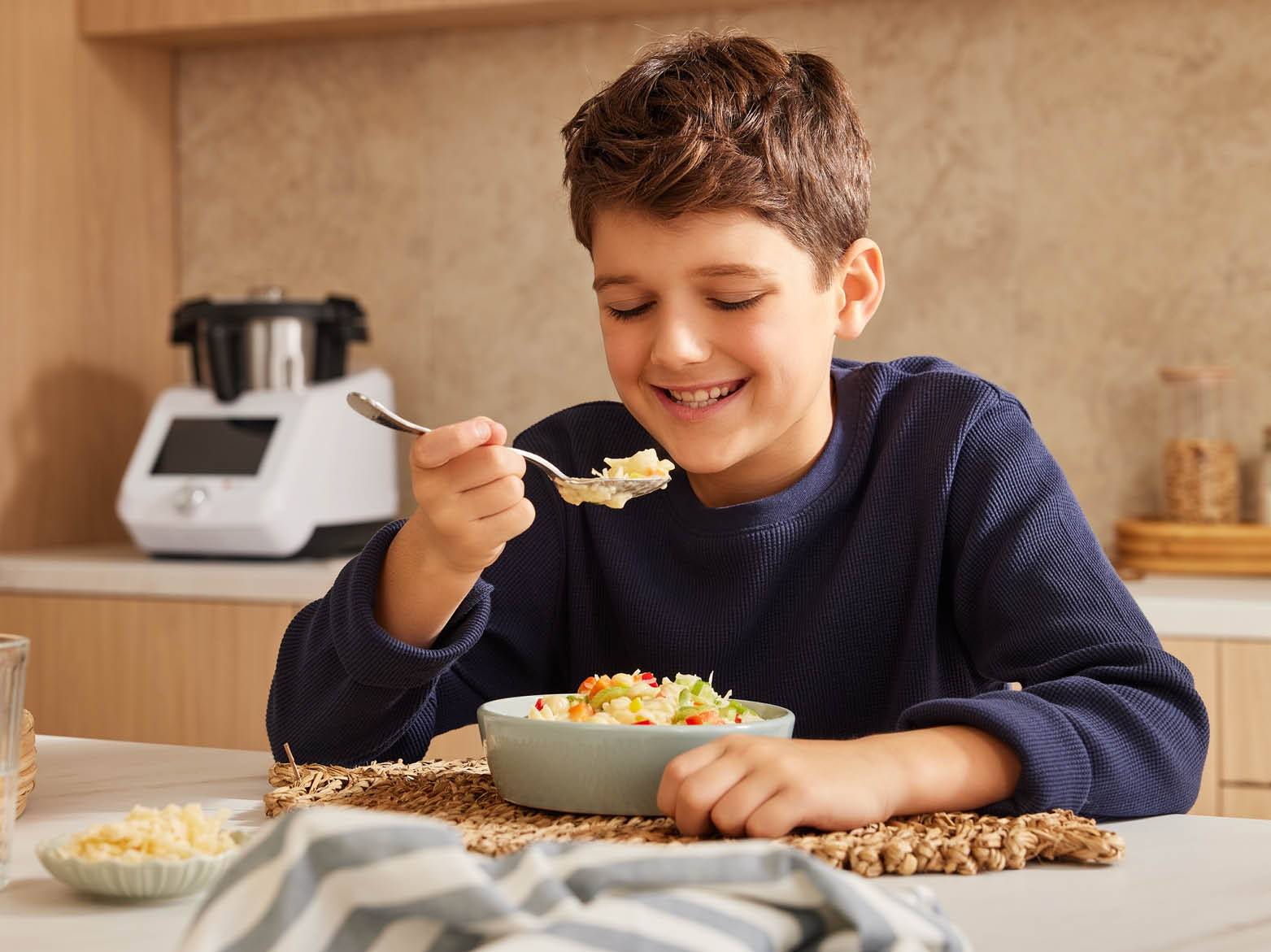 Fiú eszik tésztát egy konyhában, háttérben egy konyhai robotgéppel.