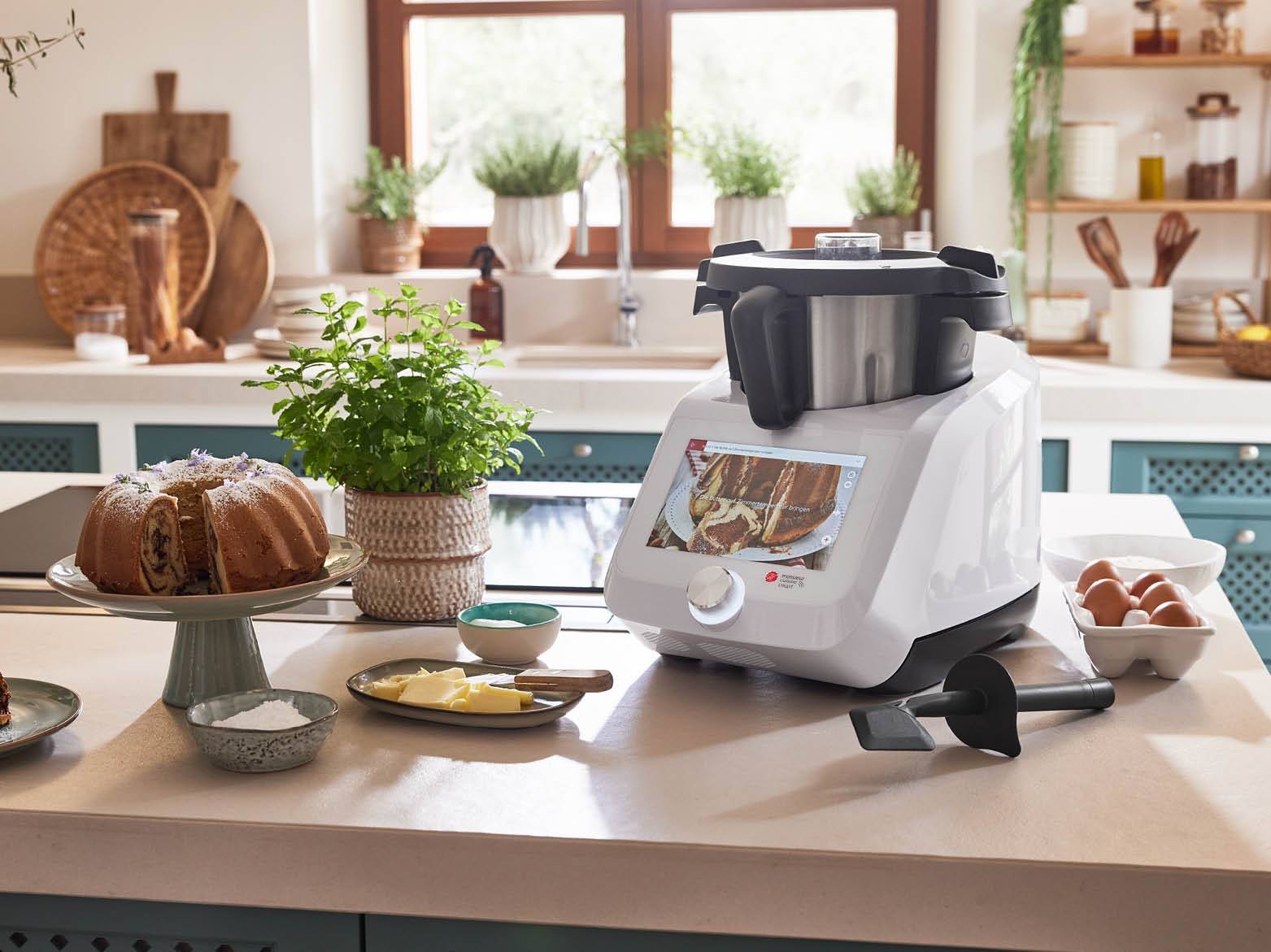 Konyhai robotgép süteménnyel és hozzávalókkal egy világos konyhában.