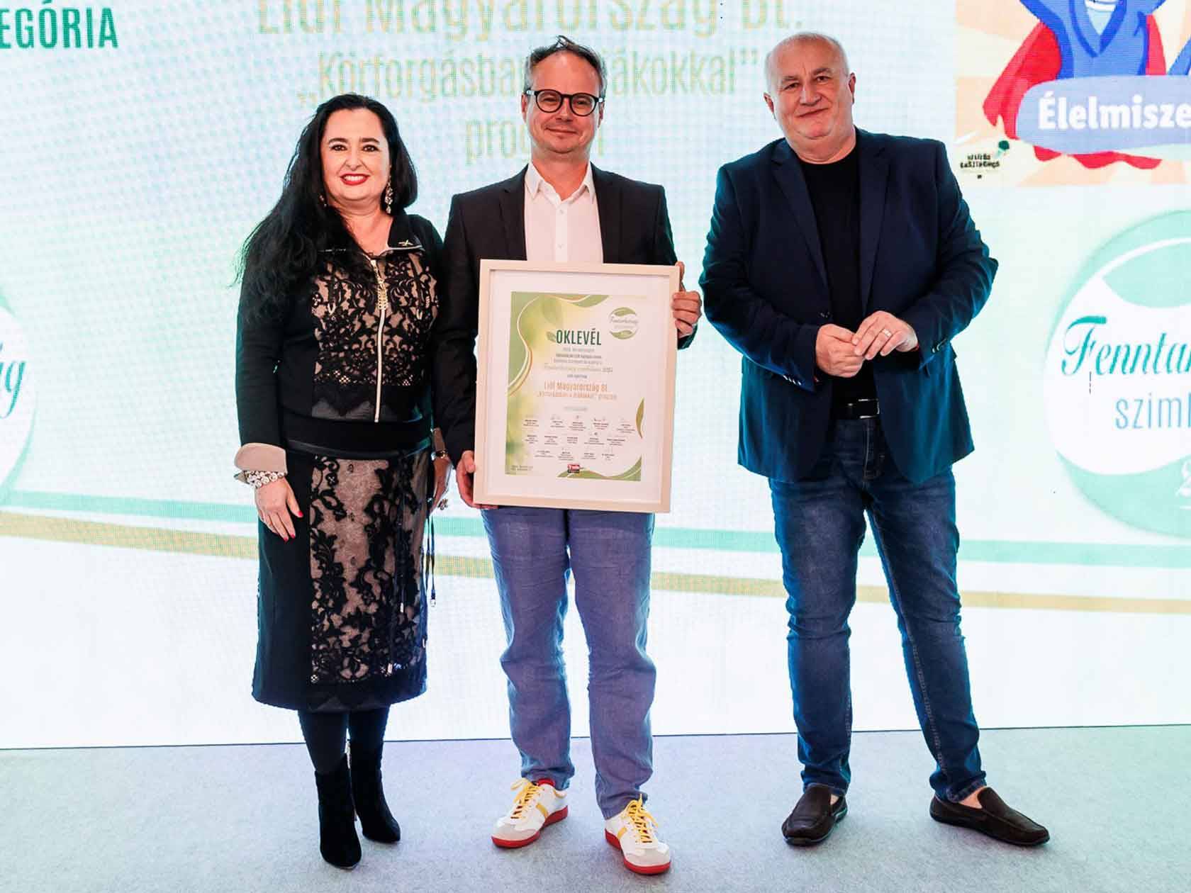 Három ember áll egy színpadon, a középső egy oklevelet tart, a háttérben a Lidl logója látható.