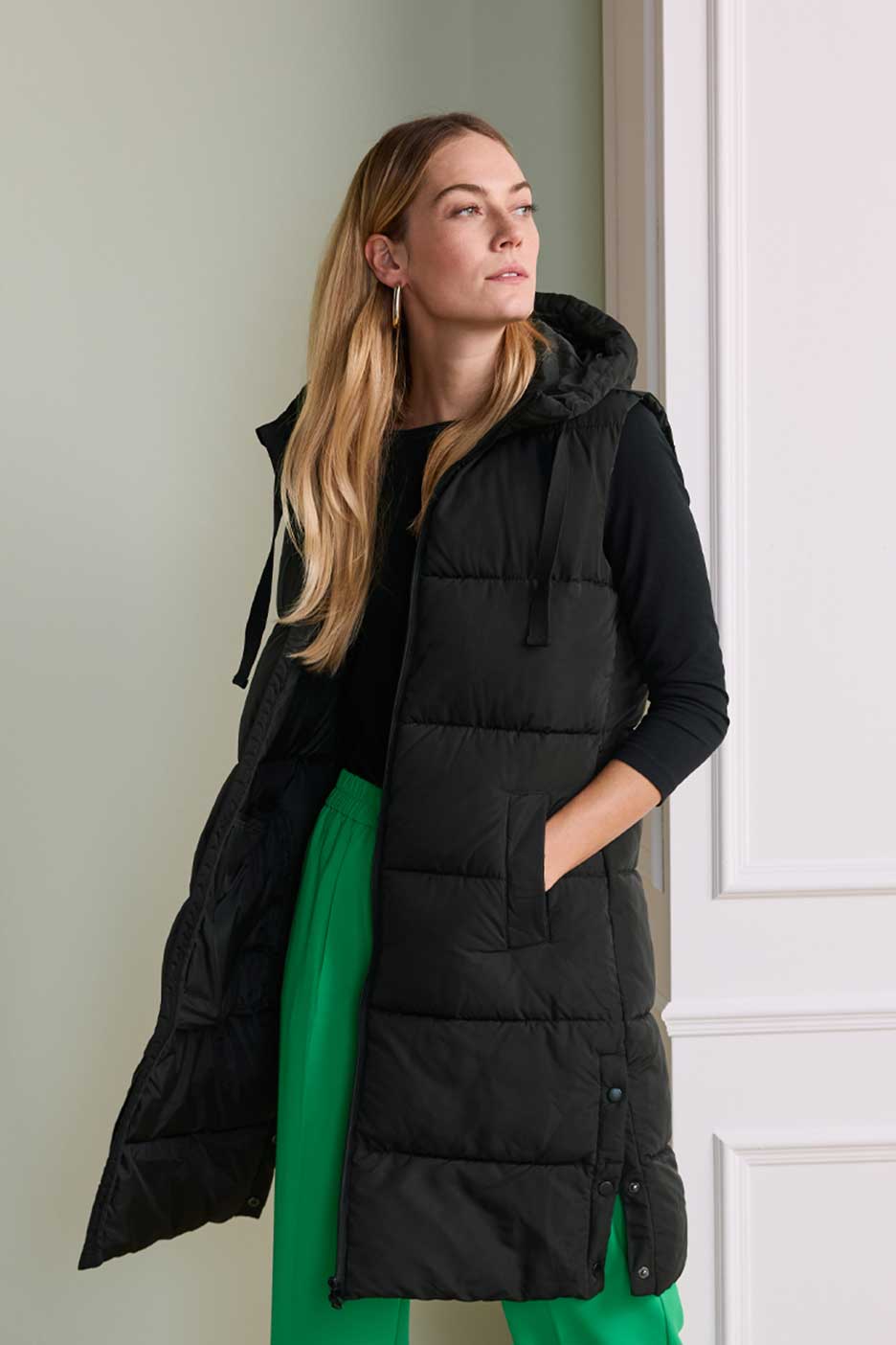 Nő hosszú fekete mellényben, kapucnival és zöld nadrággal, oldalra nézve.