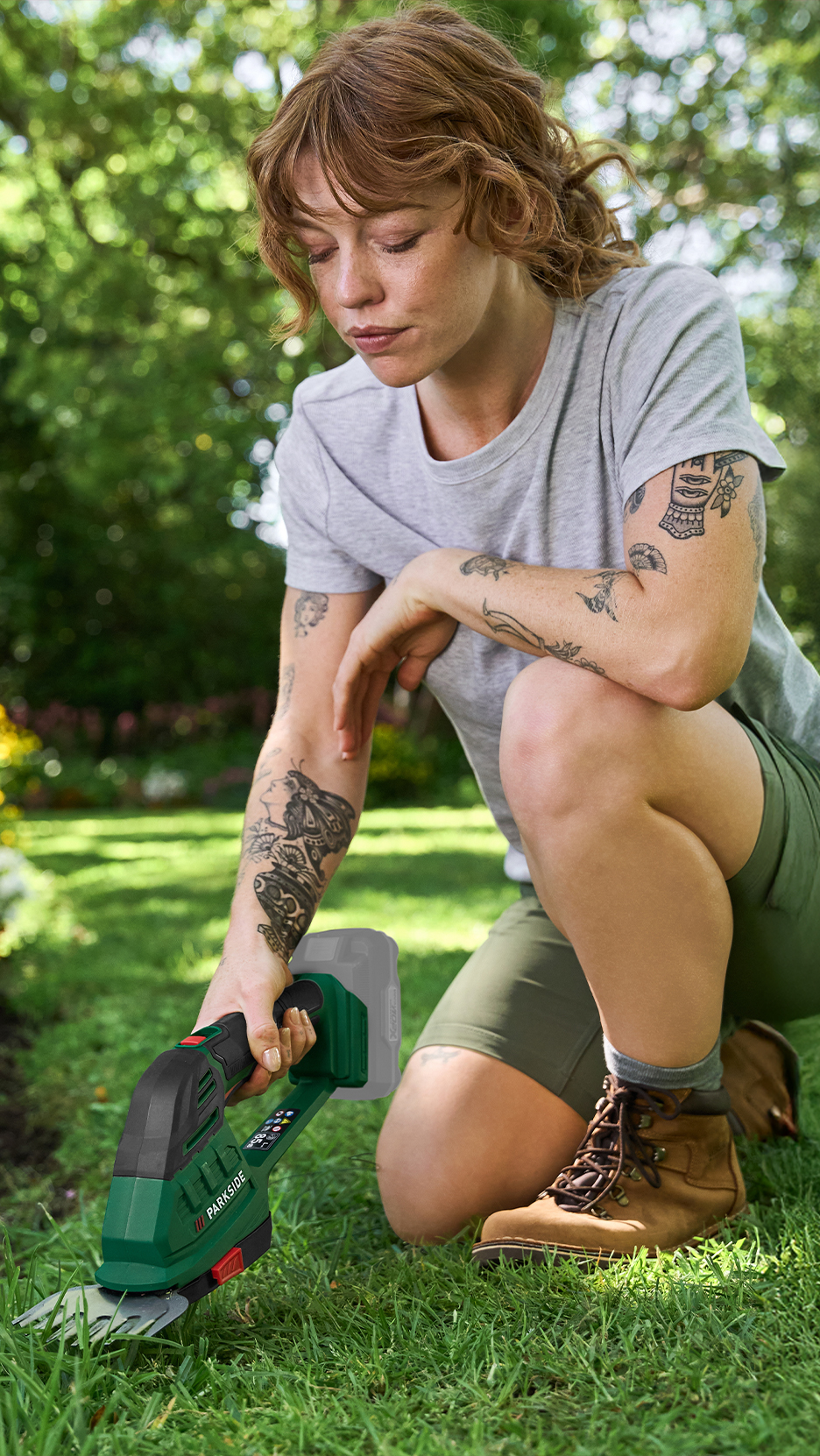 Nő térdel a füvön, akkus fűnyíróval vágja a füvet.