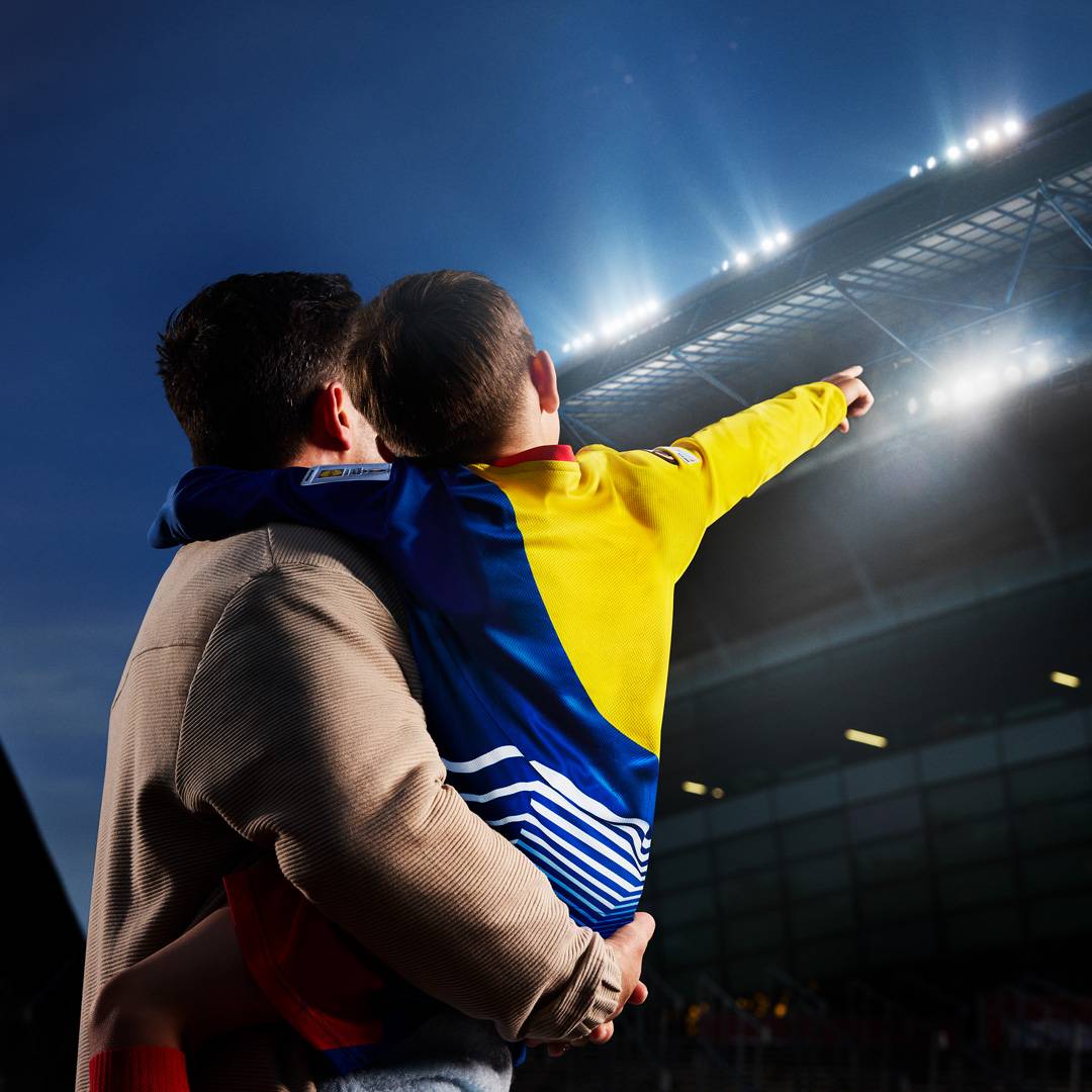 Apa és fia focimeccsen, a fiú sárga-kék mezben, apja karjában ülve mutat a pályára.