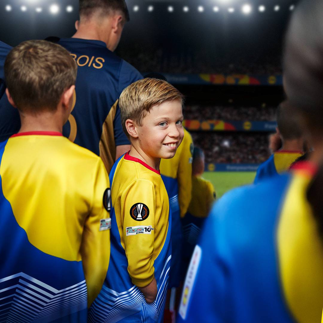 Fiúk UEFA Foundation mezben, focipályán, háttérben stadionnal.