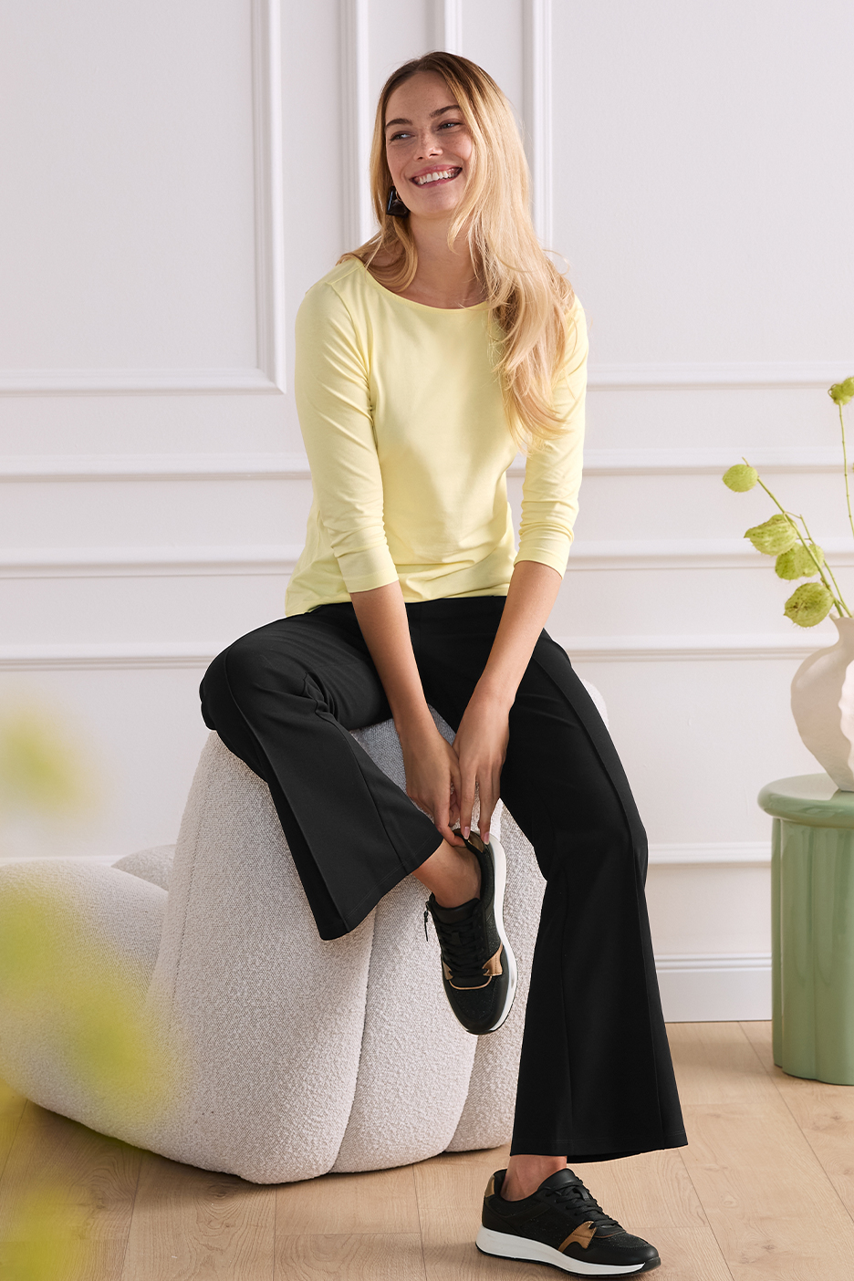 Mosolygó nő sárga hosszú ujjú felsőben és fekete nadrágban, ülve.