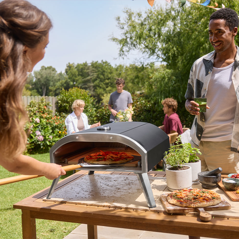 Család kerti partin, nő pizzát tesz a sütőbe, férfi italt tart.