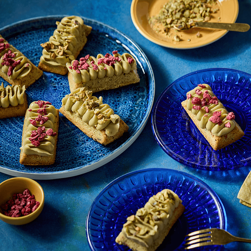 Pisztáciás és málnás financier sütemények kék tányérokon.