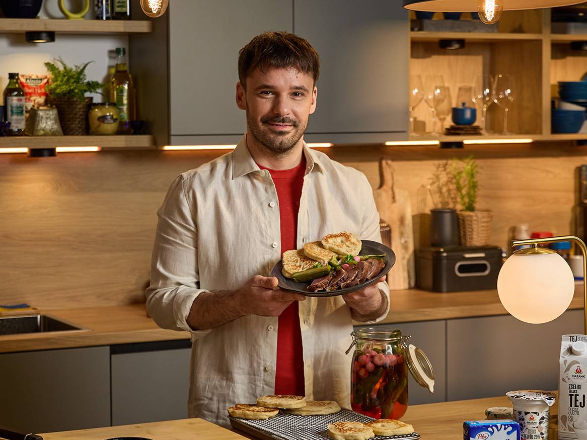 Egy férfi szakács egy tálban sült húst és pogácsát tart a kezében, Mazurek tejtermékek láthatóak a háttérben.