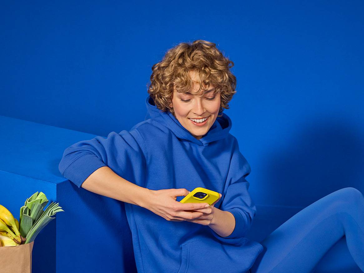 Egy fiatal nő kék pulóverben sárga telefonnal a kezében ül egy kék háttér előtt.