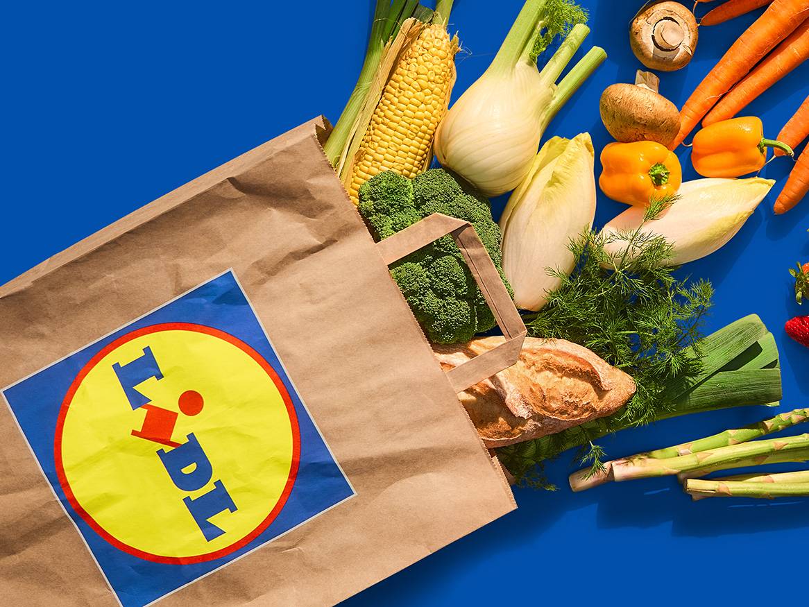 Lidl bevásárlószatyorból friss zöldségek, gyümölcsök és kenyér omlik ki egy kék háttér előtt.